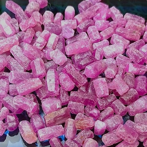 May include: A collection of raw, uncut ruby gemstones. The rubies are a vibrant pinkish-red color, with a rough, crystalline texture. The gemstones vary in size and shape, creating a natural, organic aesthetic. Ideal for jewelry making or collecting.