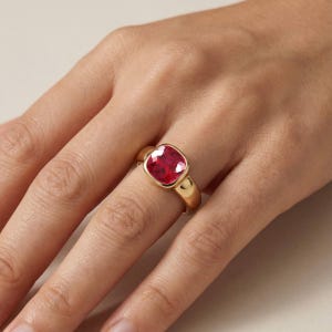 Op de afbeelding: Een gouden ring met een grote, kussengeslepen, levendige roze-rode edelsteen in een gladde, gepolijste gouden band. De gefacetteerde steen zit stevig vast in een gladde zetting en vormt een opvallend en reflecterend middelpunt op de vinger.