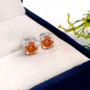 May include: A pair of stud earrings with round, orange gemstones set in silver-colored metal. The earrings are displayed inside a blue jewelry box with a white interior, showcasing the jewelry's vibrant color and design.