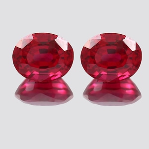 May include: Two oval-cut, vibrant red gemstones with a reflective surface beneath each. The gems are a deep, rich red color, with facets catching the light. The overall presentation suggests a focus on precious stones.