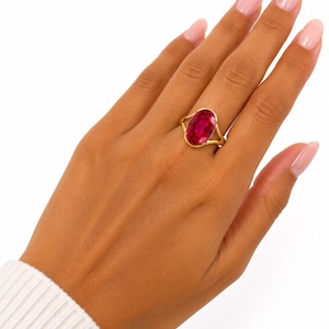 Può includere: Un anello in oro con una grande pietra preziosa rosso vivo dal taglio ovale, incastonata in una semplice fascia in oro lucido. L'anello è indossato su un dito, mettendo in risalto il colore cremisi intenso della pietra e la finitura liscia.