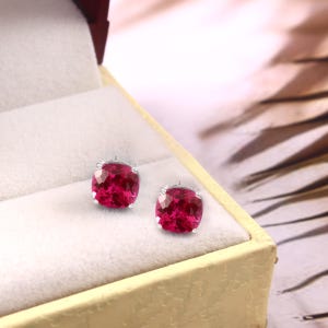 Puede incluir: Un par de pendientes de botón de piedras preciosas rojas vibrantes, de corte cuadrado, con engaste plateado, expuestos dentro de un joyero color crema. Un accesorio llamativo.