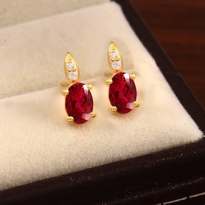 Può includere: Un paio di orecchini color oro con pietre preziose ovali rosso scuro. Ogni orecchino ha una piccola parte superiore allungata con tre piccole pietre trasparenti e un'incastonatura dorata decorativa.
