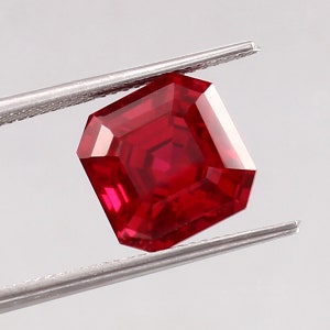 May include: A deep red gemstone with a square emerald cut. The gemstone is held in tweezers against a white background.