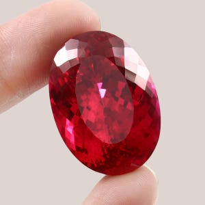 Puede incluir: Una gran piedra preciosa roja de forma ovalada con un corte facetado. La piedra preciosa se sostiene en la mano de una persona.