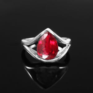 May include: A silver ring featuring a pear-shaped, faceted red gemstone set in a split-shank band. The vibrant red stone is positioned centrally within the polished metal frame, which curves upwards to a point above the gemstone's rounded base.