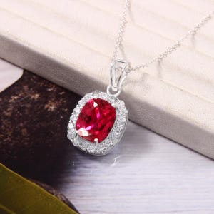 May include: A silver pendant necklace with a vibrant, oval-cut, ruby-red gemstone, encircled by a halo of small, clear crystals. The pendant is suspended from a fine silver chain, offering a timeless and sophisticated jewellery design.