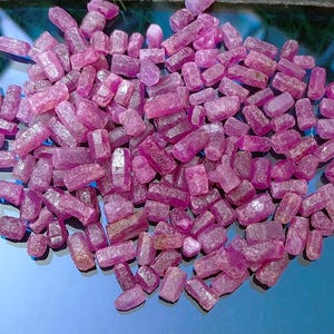 Pode incluir: Uma coleção de cristais de rubi brutos e não lapidados. Os cristais são de cor rosa-avermelhada intensa e variam em tamanho e forma. Estão espalhados sobre uma superfície azul reflexiva. Ideais para joalheria ou colecionismo.