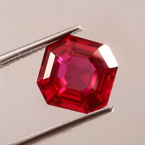 May include: A deep red octagon-shaped gemstone with a faceted cut. The gemstone is held by a pair of tweezers.