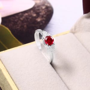 Könnte beinhalten: Ein silberfarbener Ring mit einem runden, leuchtend roten Mittelstein, der auf jeder Seite von drei kleinen, klaren, funkelnden Steinen flankiert wird und in einer hellen Schmuckschatulle aufrecht in einer polierten Metallschiene steht.
