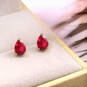 Peut inclure: Une paire de boucles d'oreilles à clous avec des pierres précieuses rouges en forme de poire, serties dans un métal doré. Chaque boucle est fixée par un sertissage à griffes, révélant une surface facettée qui reflète la lumière, dans un écrin.