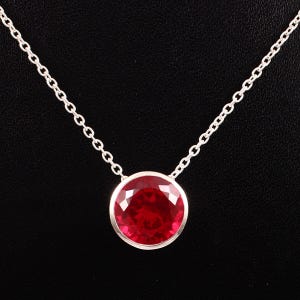 May include: A silver necklace with a large, round, faceted ruby gemstone pendant. The ruby is a vibrant red colour, set in a silver bezel, and hangs from a delicate silver chain. The background is black.