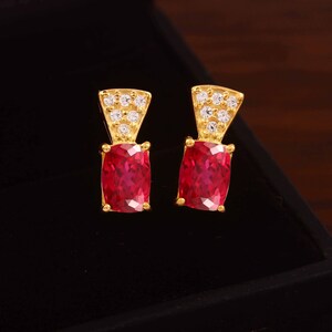 May include: A pair of gold stud earrings featuring a rectangular, faceted red gemstone at the base and a triangular top section embellished with six small, clear, round-cut white stones arranged in a cluster.