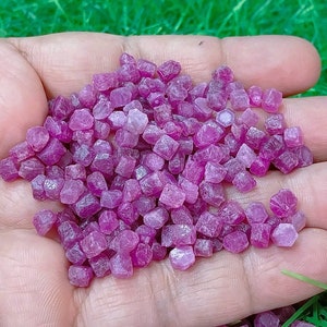 Puede incluir: Un puñado de pequeños cristales de rubí rosa. Los cristales son facetados y tienen un aspecto brillante y translúcido.