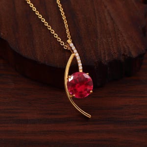 Peut inclure: Un collier en or avec un pendentif en pierre précieuse rouge vif de forme ronde, monté sur une monture dorée incurvée. La partie supérieure de la monture est ornée d'une rangée de petites pierres claires étincelantes. Chaîne dorée.