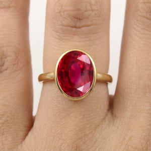 Puede incluir: Un anillo de oro con una gran piedra preciosa de color rojo intenso y corte ovalado, montada en un engaste de bisel liso y pulido. La banda es fina y circular, hecha de un metal amarillo de tonos cálidos que resalta la piedra roja facetada.