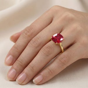 Peut inclure: Une bague en or avec une grande pierre précieuse rouge vif de taille coussin, fixée par quatre griffes. L'anneau est en or jaune poli et la pierre à facettes présente des teintes cramoisies profondes avec des reflets lumineux.