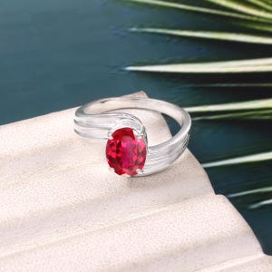 Könnte beinhalten: Ein Silberring mit einem einzelnen, oval geschliffenen, leuchtend roten Edelstein in der Mitte. Die Ringschiene besteht aus drei glatten, polierten Silbersträngen, die an beiden Seiten zusammenlaufen und den facettierten Stein halten.