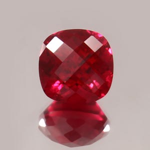 May include: A faceted, cushion-cut ruby gemstone. The gem is a deep, saturated red with brilliant internal reflections. The stone is displayed against a neutral background, with its reflection visible below.