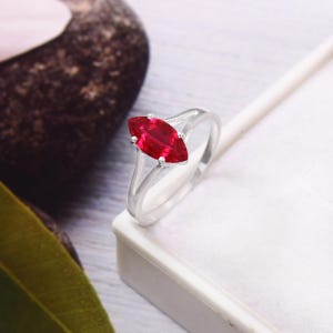 Peut inclure: Une bague en argent ornée d'une pierre rouge vif taillée en marquise. La pierre est sertie de quatre griffes, et la bague a un anneau simple et élégant. La bague est présentée sur une surface blanche.