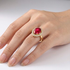 Op de afbeelding: Een gouden ring met een grote, peervormige, levendig rode edelsteen in het midden. De centrale steen is omringd door een delicate halo van kleine, heldere, sprankelende witte steentjes die in de gepolijste gouden band zijn gezet.
