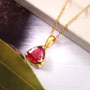 Peut inclure: Un collier en or avec un pendentif en pierre précieuse rouge profond taillée en triangle. La pierre rouge à facettes est sertie dans une monture en métal doré poli avec une chaîne assortie, offrant une couleur vive et une forme triangulaire.