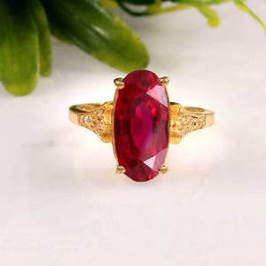 Könnte beinhalten: Ein Goldring mit einem großen, oval geschliffenen, tiefroten Edelstein, der von vier Krappen gehalten wird. Die Ringschiene besteht aus Gelbgold und ist an den Schultern beidseitig mit kleinen, klaren, im Pavé-Stil gefassten Steinen besetzt.