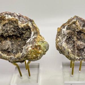 Puede incluir: Dos especímenes de geodas con un exterior rugoso marrón y un interior hueco forrado con cristales blancos. Las geodas se muestran en soportes de acrílico transparente con varillas de metal dorado.