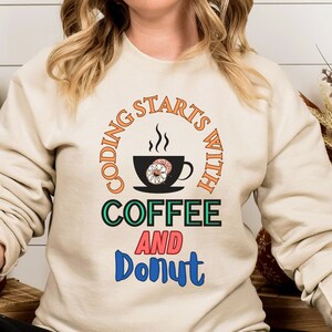 May include: A beige sweatshirt with a black circle graphic that says "Coding starts with coffee and donut". The graphic features a steaming cup of coffee with a donut inside and a donut below the text.