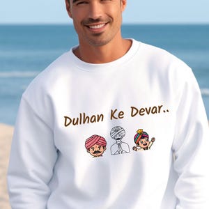 May include: White sweatshirt with the text "Dulhan Ke Devar.." in brown. The sweatshirt features three cartoon illustrations of men wearing turbans in various colors. The background is a blurred image of the ocean and sky.