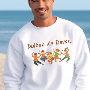 May include: White sweatshirt with the text "Dulhan Ke Devar.." in brown. The sweatshirt features a colorful illustration of dancing figures in traditional attire, holding sticks. The figures are in shades of orange, blue, green, and yellow.