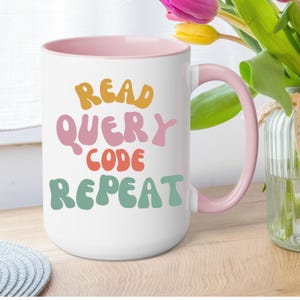 May include: White ceramic mug with a pink interior and handle. The mug features the words "READ QUERY CODE REPEAT" in colorful, retro-style lettering. The mug is on a wooden surface with flowers in the background.