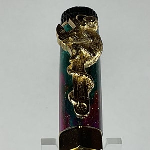 May include: A green and pink pen with a gold monkey figurine on top. The monkey is holding onto a branch and has a detailed design.