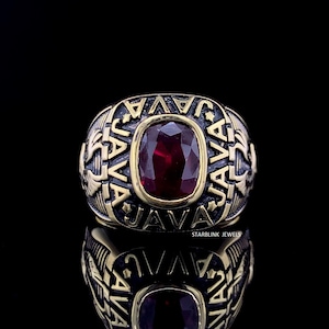 Peut inclure: Bague dorée avec une grosse pierre rouge foncé ovale au centre. Le mot "JAVA" est gravé autour de la pierre. La bague a un style vintage, avec des détails complexes.