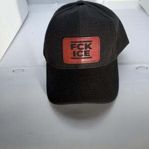 Puede incluir: Gorra de béisbol negra con visera curva. Un parche rectangular rojo en la parte delantera muestra el texto "FCK ICE" en letras negras. La gorra está hecha de tela texturizada.