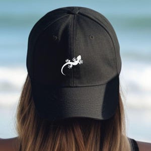 Può includere: Cappellino da baseball nero con un disegno di lucertola bianca sul davanti. Il cappellino è realizzato in un materiale morbido e ha una visiera curva. Il disegno della lucertola è un'illustrazione semplice e stilizzata.