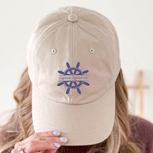 Könnte beinhalten: Beige Baseballkappe mit gebogenem Schirm. Die Kappe zeigt ein blaues nautisches Steuerraddesign mit dem Text "Captain Sparrow" darüber und darunter. Hergestellt aus einem weichen Stoff.