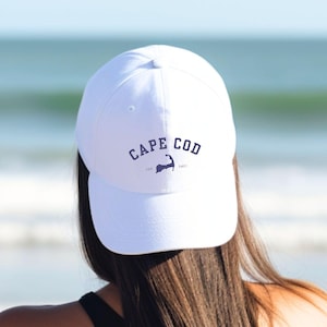 Könnte beinhalten: Weiße Baseballkappe mit dem marineblauen Schriftzug "CAPE COD" und einer marineblauen Wal-Illustration. Die Kappe wird von einer Person mit Blick auf das Meer getragen.