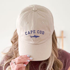 Könnte beinhalten: Beige Baseball Cap mit dem Schriftzug "CAPE COD" in Marineblau und einer marineblauen Hai-Grafik. Die Kappe besteht aus einem weichen Material und hat einen gebogenen Schirm. Ein lässiges Accessoire.