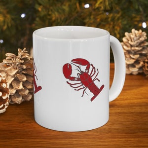Peut inclure: Une tasse en céramique blanche avec un motif de homard rouge. Le homard est dessiné dans un style simple et cartoon.