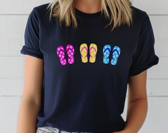 May include: Navy blue t-shirt featuring a row of colorful flip-flops. The flip-flops are in shades of pink, yellow, and blue, each adorned with a floral pattern. A casual and summery design.