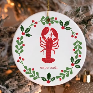 May include: A round white Christmas ornament featuring a red lobster illustration in the center, surrounded by a green and red holly wreath. The words "cape cod." are printed below the lobster. A gold string is attached for hanging.