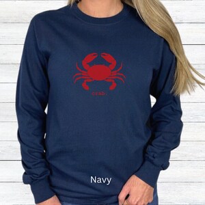 A woman wearing a Navy unisex long sleeve t-shirt with a graphic of a red crab, above the word crab in lower case font, followed by a period.