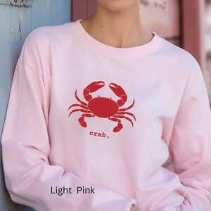 A woman wearing a Light Pink unisex long sleeve t-shirt with a graphic of a red crab, above the word crab in lower case font, followed by a period.