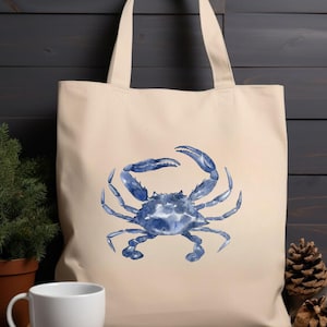 May include: A beige tote bag featuring a watercolor illustration of a blue crab. The crab is centered on the bag, with its claws raised. The bag has two handles and is made of a sturdy fabric, perfect for carrying groceries or other items.