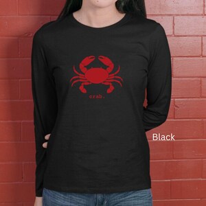 A woman wearing a black unisex long sleeve t-shirt with a graphic of a red crab, above the word crab in lower case font, followed by a period.