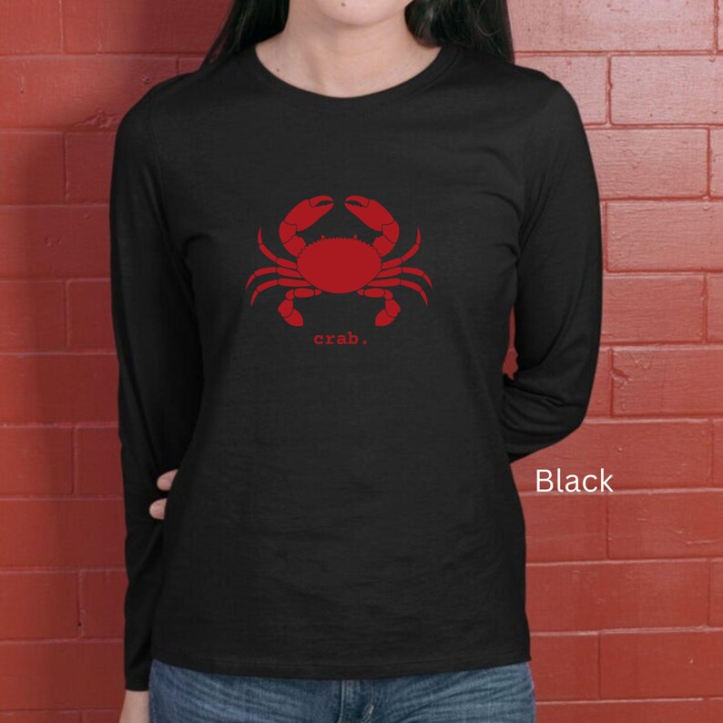 A woman wearing a black unisex long sleeve t-shirt with a graphic of a red crab, above the word crab in lower case font, followed by a period.