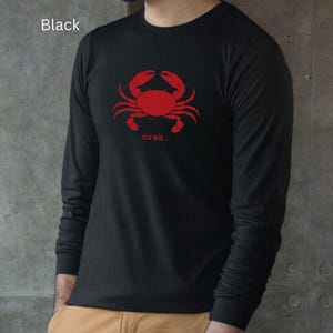 A man wearing a black unisex long sleeve t-shirt with a graphic of a red crab, above the word crab in lower case font, followed by a period.
