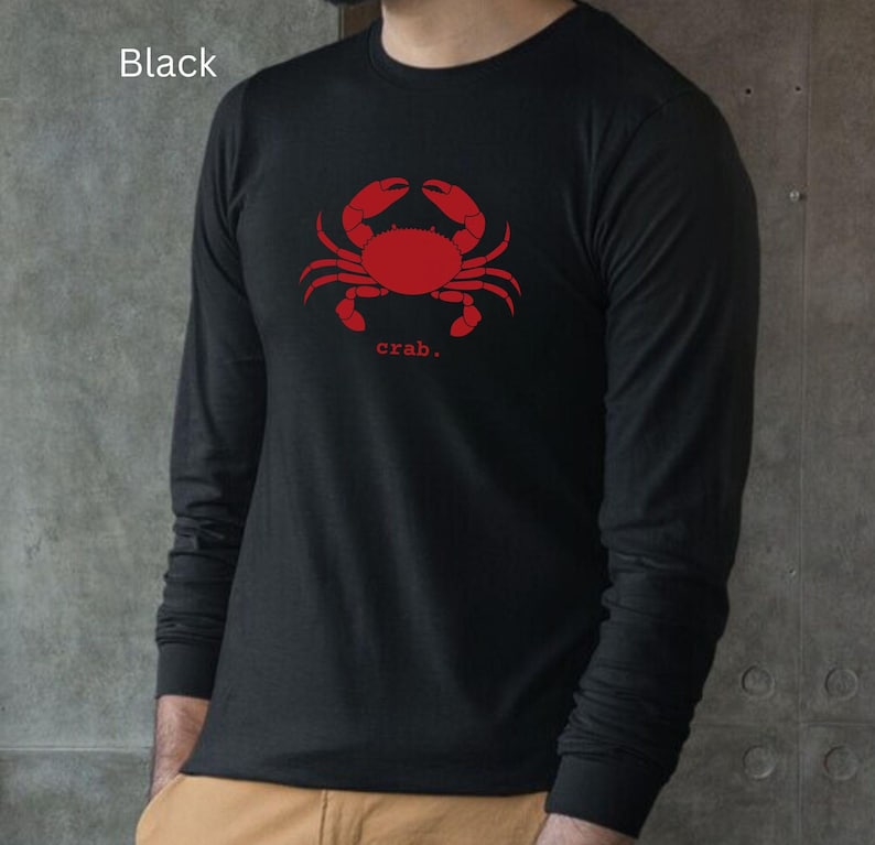 A man wearing a black unisex long sleeve t-shirt with a graphic of a red crab, above the word crab in lower case font, followed by a period.