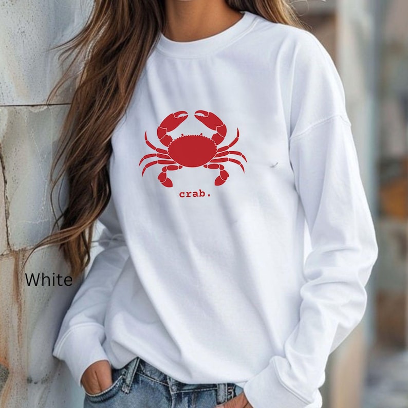 A woman wearing a White unisex long sleeve t-shirt with a graphic of a red crab, above the word crab in lower case font, followed by a period.
